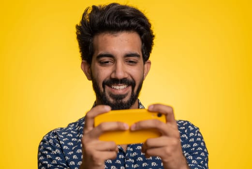 Worried funny addicted indian man enthusiastically playing racing video games on mobile phone. Handsome young guy using smartphone gadget app with drive simulator isolated on yellow studio background