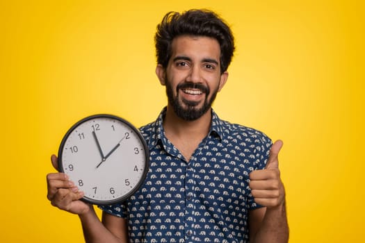 It is your time. Excited happy young indian man showing time on wall office clock, ok, thumb up, approve, pointing finger at camera. Handsome hindu guy isolated on studio yellow background indoors