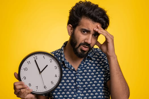 Young indian man with anxiety checking time on clock, running late to work, being in delay, deadline. Hindu guy looking at hour, minutes, worrying to be punctual isolated on studio yellow background