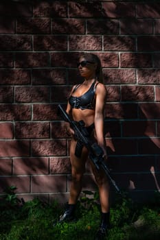 A caucasian woman in a bikini holds a machine gun against a brick wall