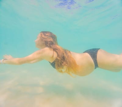 Pregnant woman underwater swimming in tropical sea. Healthy and active pregnancy. Young expecting mom on summer beach vacation before baby birth. Swim holiday and water fun. Travel during pregnancy. Underwater photography.