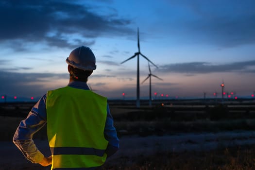 Rear view of maintenance worker wearing helmet and vest checking in wind turbine farm at sunset. Copy space. Renewable energies concept.