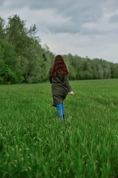 a woman in a long raincoat runs across a field in tall green grass in cloudy weather in spring. High quality photo