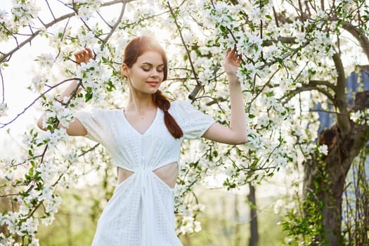 a slender, happy woman in a light dress poses next to a flowering tree in the countryside. High quality photo