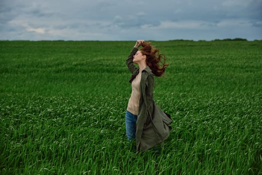 a woman in a dark coat stands in a green field against a cloudy sky holding her hair in the wind. High quality photo