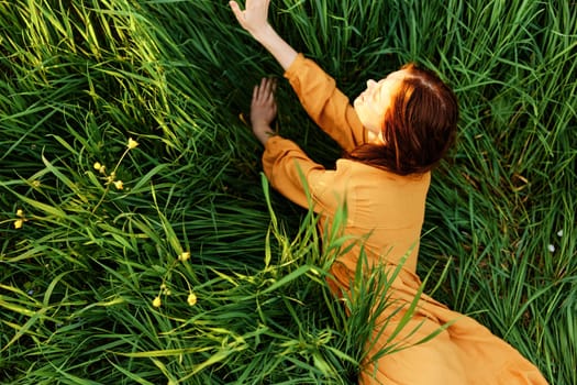 a relaxed red-haired woman enjoys summer lying in the tall green grass in a long orange dress smiling happily looking away. High quality photo