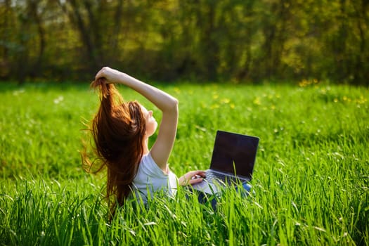 a red-haired woman sits in a field of green grass with a laptop and straightens her long hair. High quality photo