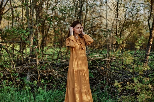 a beautiful woman walks in the shade near the trees, dressed in a long orange dress, enjoying the weather and the weekend, straightening her hair with her hands. The theme of privacy with nature,. High quality photo