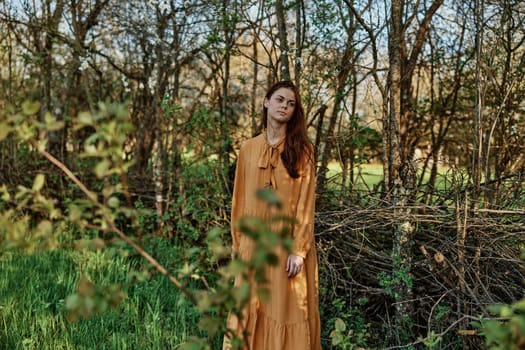 a beautiful, slender woman with long hair walks in the shade near the trees, dressed in a long orange dress, enjoying the weather and the weekend. The theme of privacy with nature. High quality photo