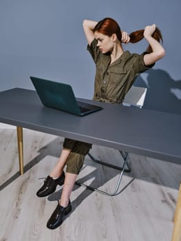 a woman in overalls sits at a laptop in the office and collects her hair with her hands. High quality photo