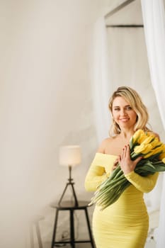 a happy woman in a yellow dress embraces a bouquet of yellow spring tulips in the interior.