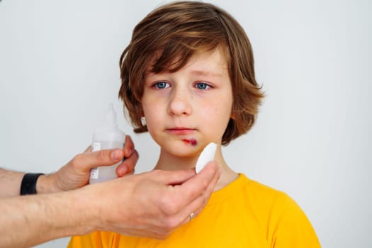 Dad doctor father treats bruised wound on his son school boy kid face. Man cleans addresses the sore wound on child face on white background with copy space for text.