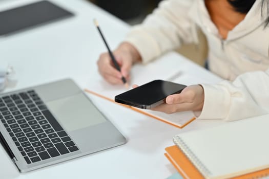 Cropped image of student woman holding smart phone and writing on notepad at working desk.