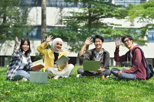 Group of young students sitting outside of university building and waving hands to camera. Youth lifestyle and education concept.
