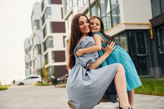 Embracing each other. Young mother with her little daughter walking near the buildings.