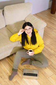 Young Asian woman wearing glasses is sitting comfortably on the floor with laptop in front of her, holding cup of coffee. She is engaged in digital work, enjoying moment of relaxation with her favorite beverage while working from home. High quality photo