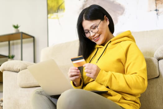 Asian woman is comfortably shopping online from the comfort of her home, with laptop on her lap and bank card in her hand. She is engaged in digital retail, using modern technology and the convenience of e-commerce to make purchase with her credit card. High quality photo