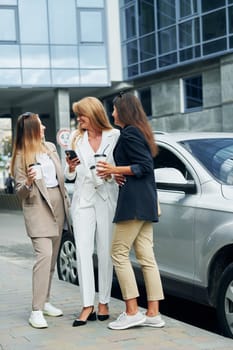 Women in formal wear is outdoors in the city together standing near silver colored car.