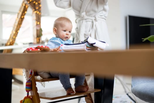 Happy infant sitting at dining table and playing with his toy in traditional scandinavian designer wooden high chair in modern bright atic home superwised by his mother.