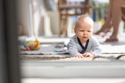 Cute little infant baby boy playing with toys outdoors at the patio in summer being supervised by her mother seen in the background. Selective focus.