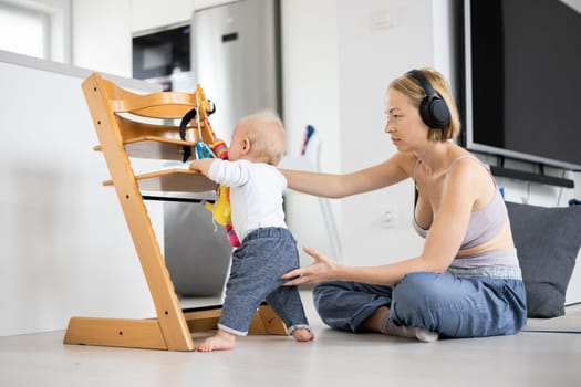 Women's multitasking. Mother sitting on floor playing with her baby boy watching and suppervising his first steps while listening to podcast on wireless headphones