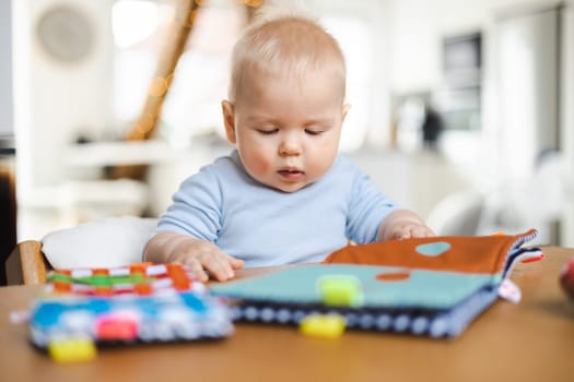 Happy infant sitting at dining table and playing with his toy in traditional scandinavian designer wooden high chair in modern bright atic home. Cute baby playing with toys.