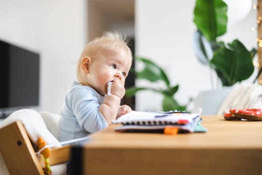 Happy infant sitting at dining table and playing with his toy in traditional scandinavian designer wooden high chair in modern bright atic home. Cute baby playing with toys.