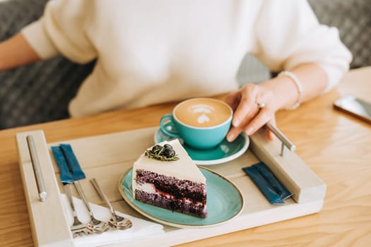 Mature woman's hands holding a cup of cappuccino coffee and a cream cake. Adult lady in white sweater enjoying her breakfast latte and dessert pie on a wooden tray in a cafe.