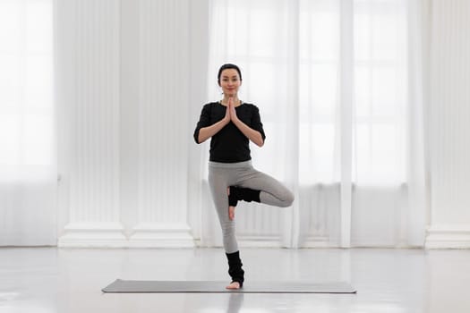 Beautiful young woman doing tree pose during yoga meditation in the white gym. The concept of awareness, calmness of the nervous system, balance and harmony with oneself.