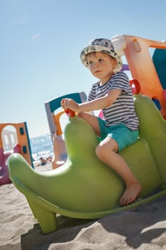 A little boy poses on a green plastic toy at a beach playground. Sand, sea and light blue sky in the background