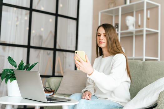 Freelance young woman with mobile cellphone typing at laptop and working from home office with. Happy girl on workplace at desk. Distance learning online education. Taking selfie or video chat