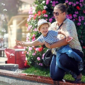 Mom holds her little boy on hand as they try to touch the water in the fountain. A beautiful city celebration on a sunny summer day.