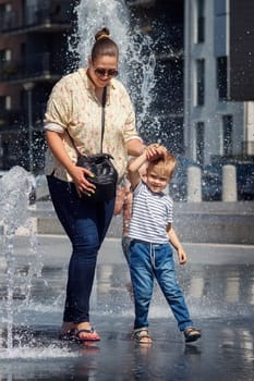 Mom and her son walk around town near the fountain on a hot summer day. High water flow, possibility to get wet, dark blue background of city buildings.