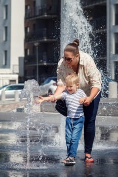 Mom and her son, in the city center, by a fountain of water. The child is afraid to touch the water so that it does not get wet, the mother encourages it.