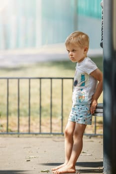 A blond, barefoot boy in a white shirt takes a break on an outdoor playground on a hot summer day.