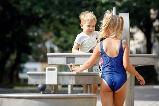 A happy little boy and nice girl in a blue swimsuit plays with a water tap in a city park. Special water equipment for children's games on a hot summer day outdoors. Horizontal photo.