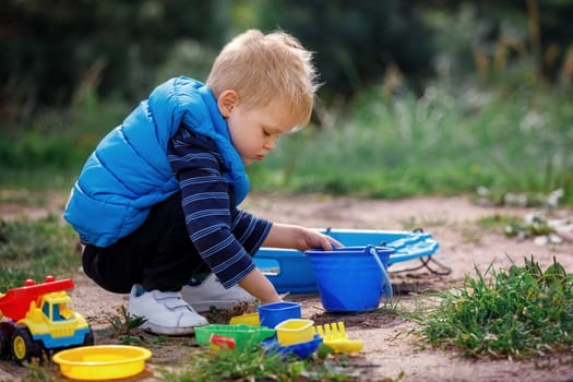 A little boy in blue clothes plays with sand in nature. The child has a beautiful colorful set of plastic beach toys.