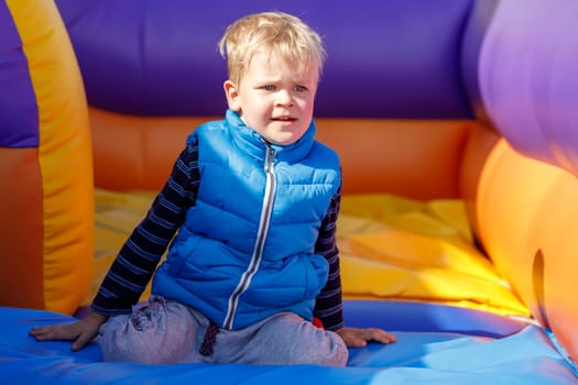 A portrait of a little boy wearing a blue vest on a colorful striped trampoline. The child squats for a little rest.