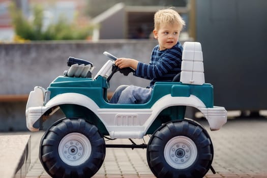 Funny boy car driver with the steering wheel. Three year-old boy in a blue sweater in a green toy car in the city. Little boy driving big toy car and having fun, outdoors.