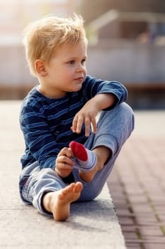 A portrait of a cute little boy sitting by a city fountain, the child takes off his socks, he wants to cool his feet in the cool water.