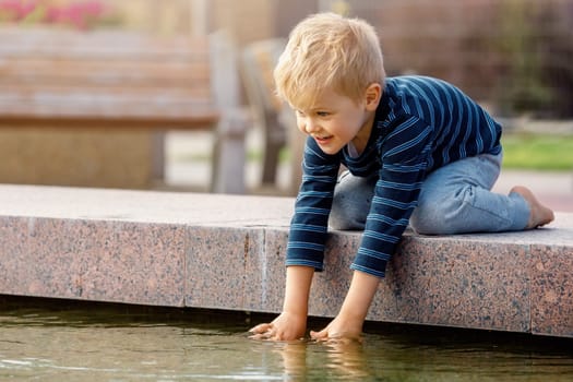 Portrait of adorable 3 years old toddler boy touching water in fountain at park.