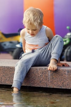 A little funny, blond boy in town tests the water by dipping one foot in a fountain.