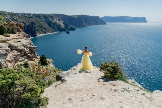 Woman in a yellow dress on the sea. Side view Young beautiful sensual woman in yellow long dress posing on a rock high above the sea at sunset. Girl in nature against the blue sky.