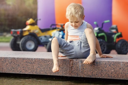 A little barefoot boy in the city plays near the fountain with water at summer.