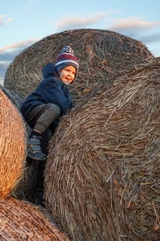 A naughty little boy climbs to the top of a bale of straw in the autumn evening in the light of sunset.