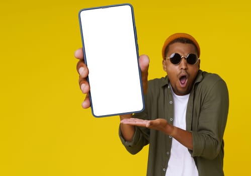 Surprised African man is holding phone with white blank screen against yellow background with copy space. The image is ideal for advertising concepts. High quality photo