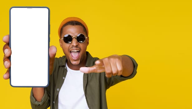 Young man is seen pointing towards camera while showcasing his smartphone with white blank screen on yellow background. Concept of communication and technology, promoting use of mobile devices and social media for business and personal purposes. High quality photo
