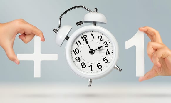 Change time. Spring forward, on a gray background. A white alarm clock indicates that the time has been moved forward an hour. Hand holding number one with plus sign, Spring forward concept.