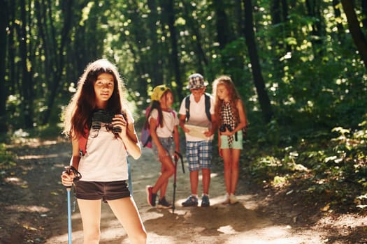 Girl standing in front of her friends. Kids strolling in the forest with travel equipment.