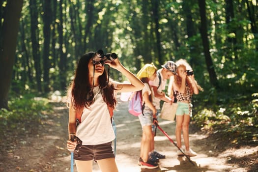 Girl standing in front of her friends. Kids strolling in the forest with travel equipment.